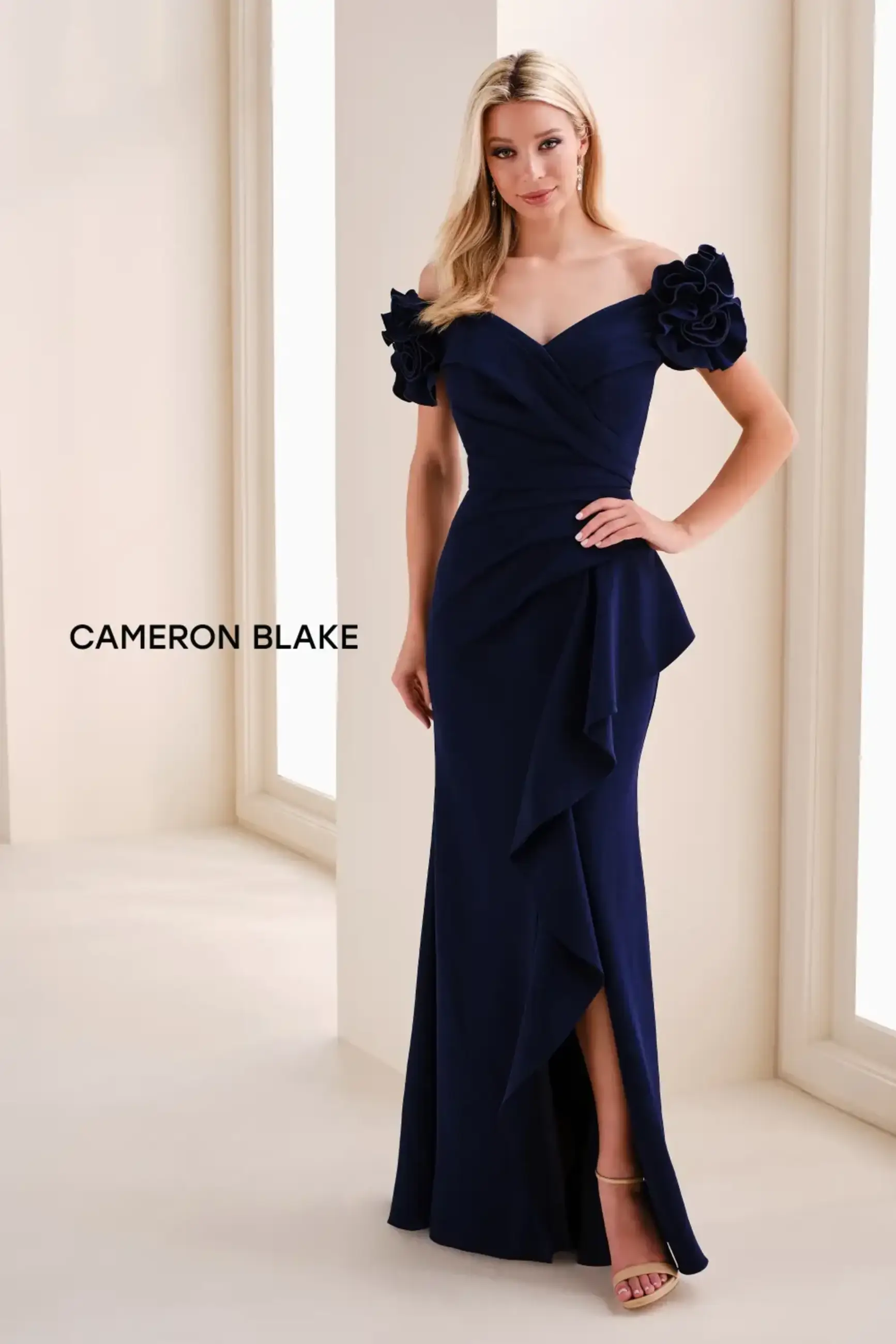 A woman in a navy blue gown with off-the-shoulder sleeves and floral accents, standing in an elegant setting. The dress features a fitted bodice and a ruffled detail down one side.