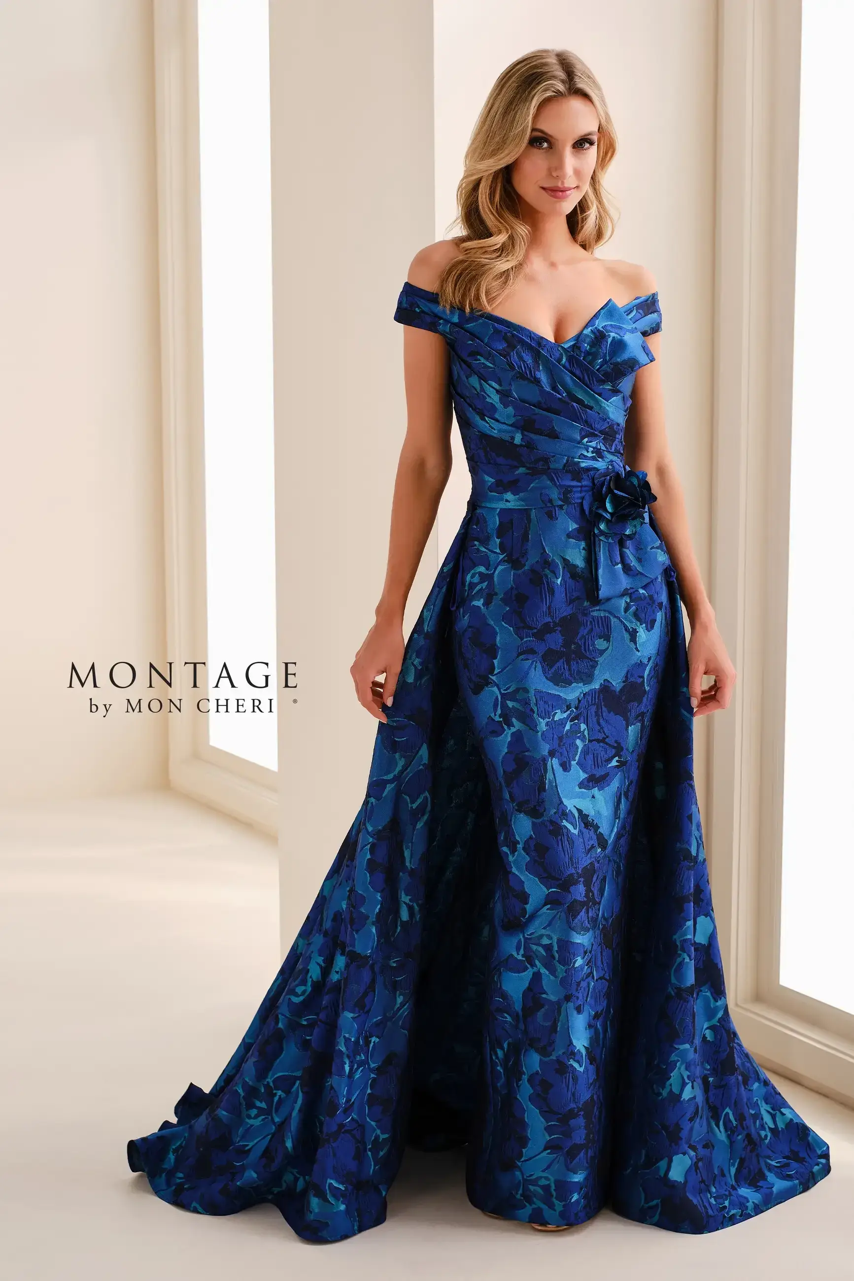 A woman in an elegant blue floral off-shoulder gown poses gracefully in a bright room. The dress features a fitted bodice and flowing skirt.