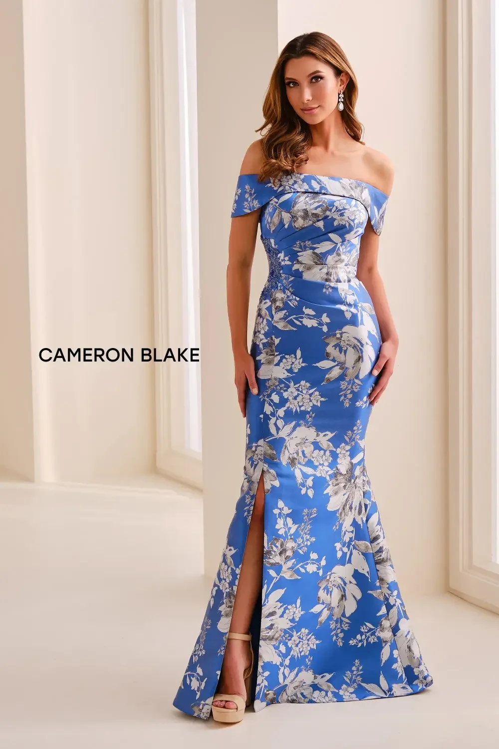 A woman in an off-shoulder blue floral dress standing in a light-colored room.