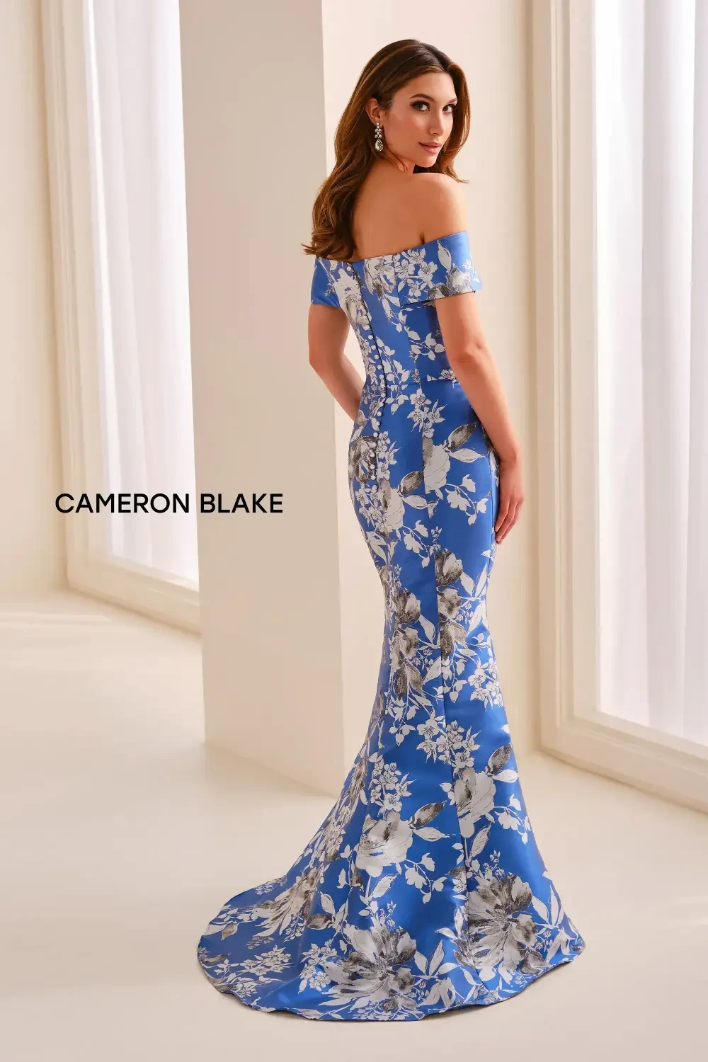 A woman in an off-shoulder blue floral dress standing in a light-colored room.