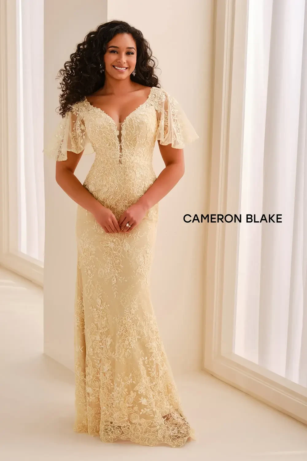 A woman in a gold-toned lace gown with short, sheer sleeves, standing in a well-lit room.