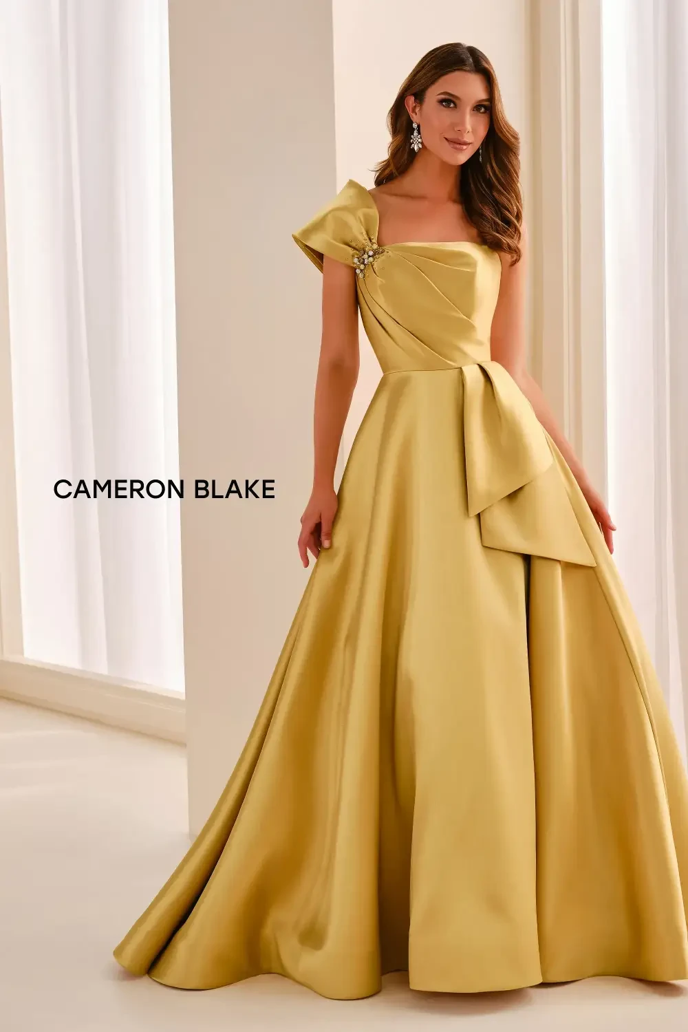 A woman wearing a yellow evening gown with one shoulder design and a bow detail, standing in a bright room.