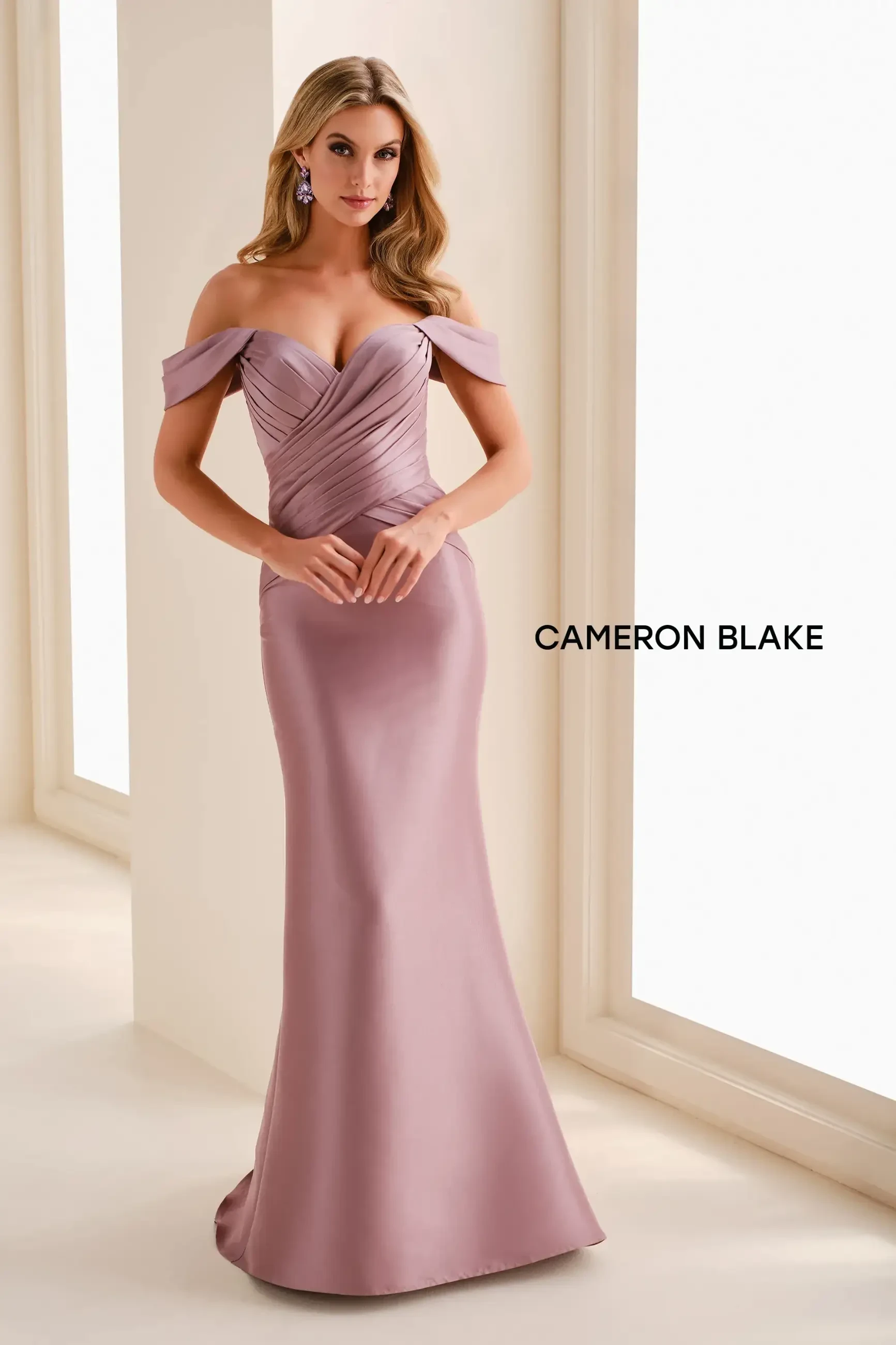 Elegant woman in a mauve off-shoulder gown stands confidently in a softly lit space. The gown features intricate pleating, evoking a graceful tone.