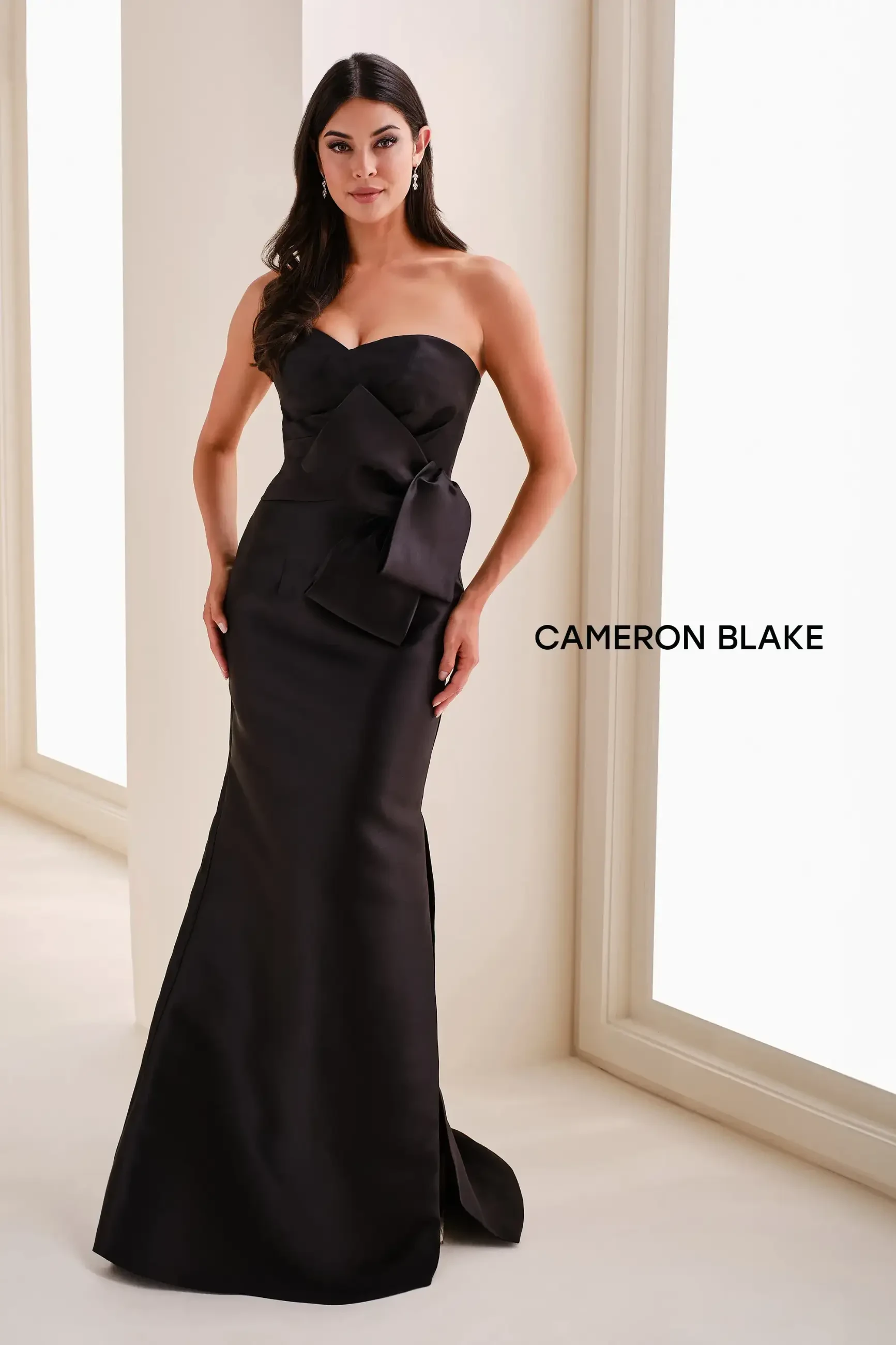 A woman in an elegant black strapless gown with a bow detail, standing in a bright room.