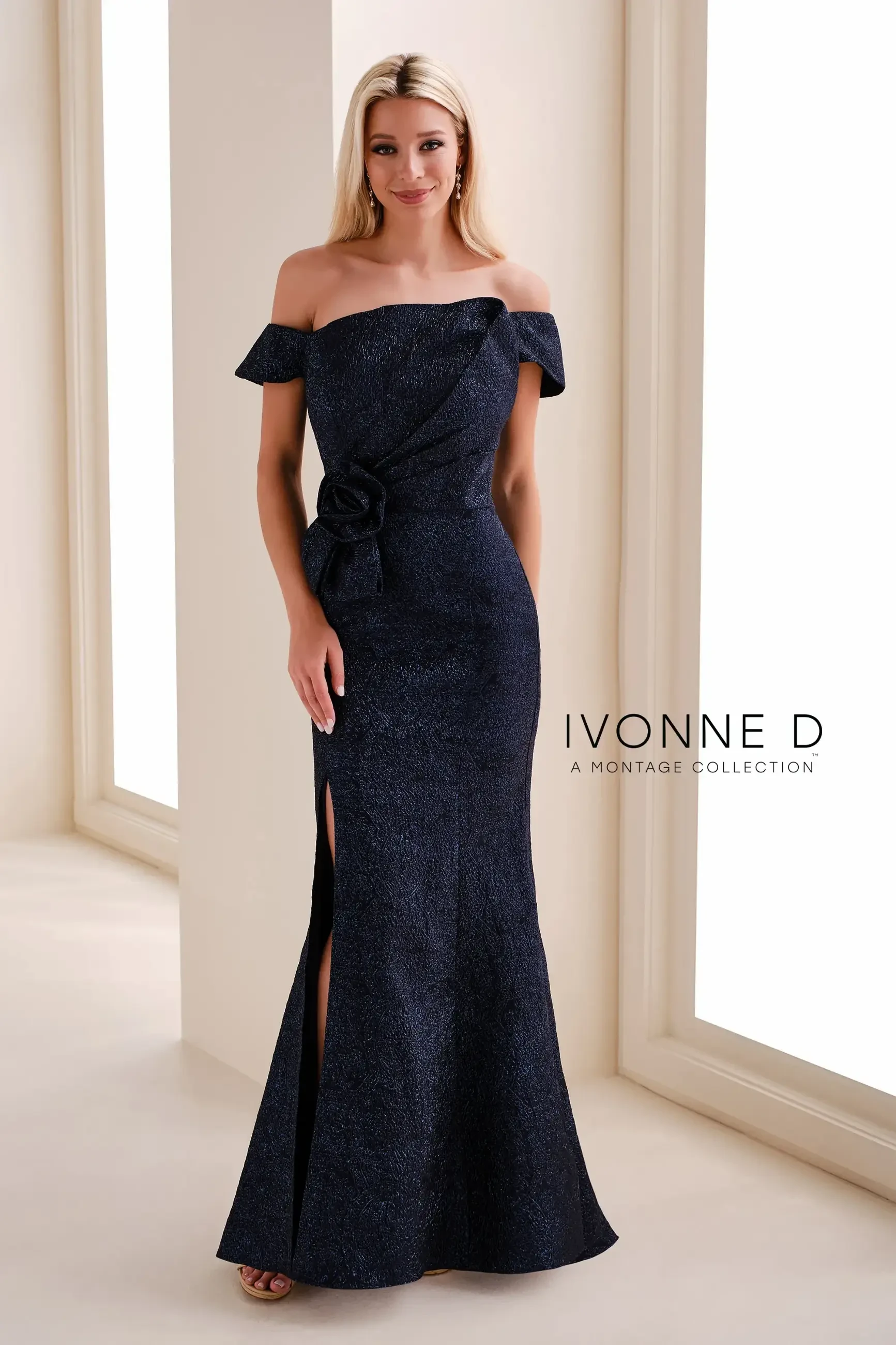 A woman in an elegant off-the-shoulder navy gown with a slit and a floral accent at the waist, standing in a softly lit space.