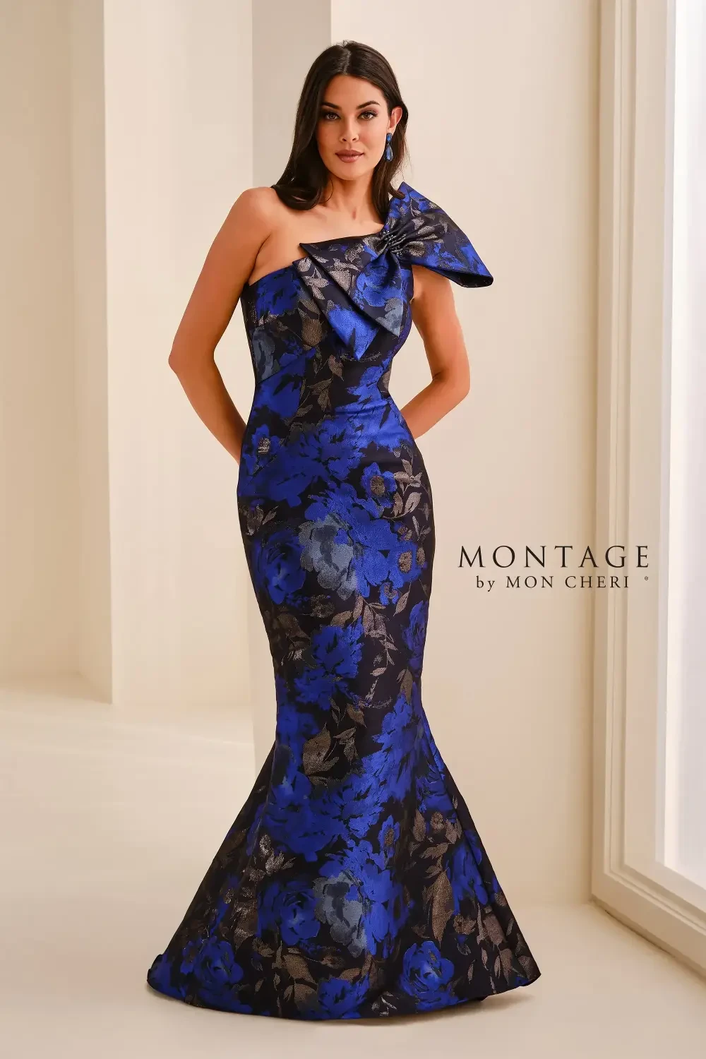 A woman wearing a blue and black floral evening gown with one shoulder and a large bow detail, posed elegantly in a light-colored room.