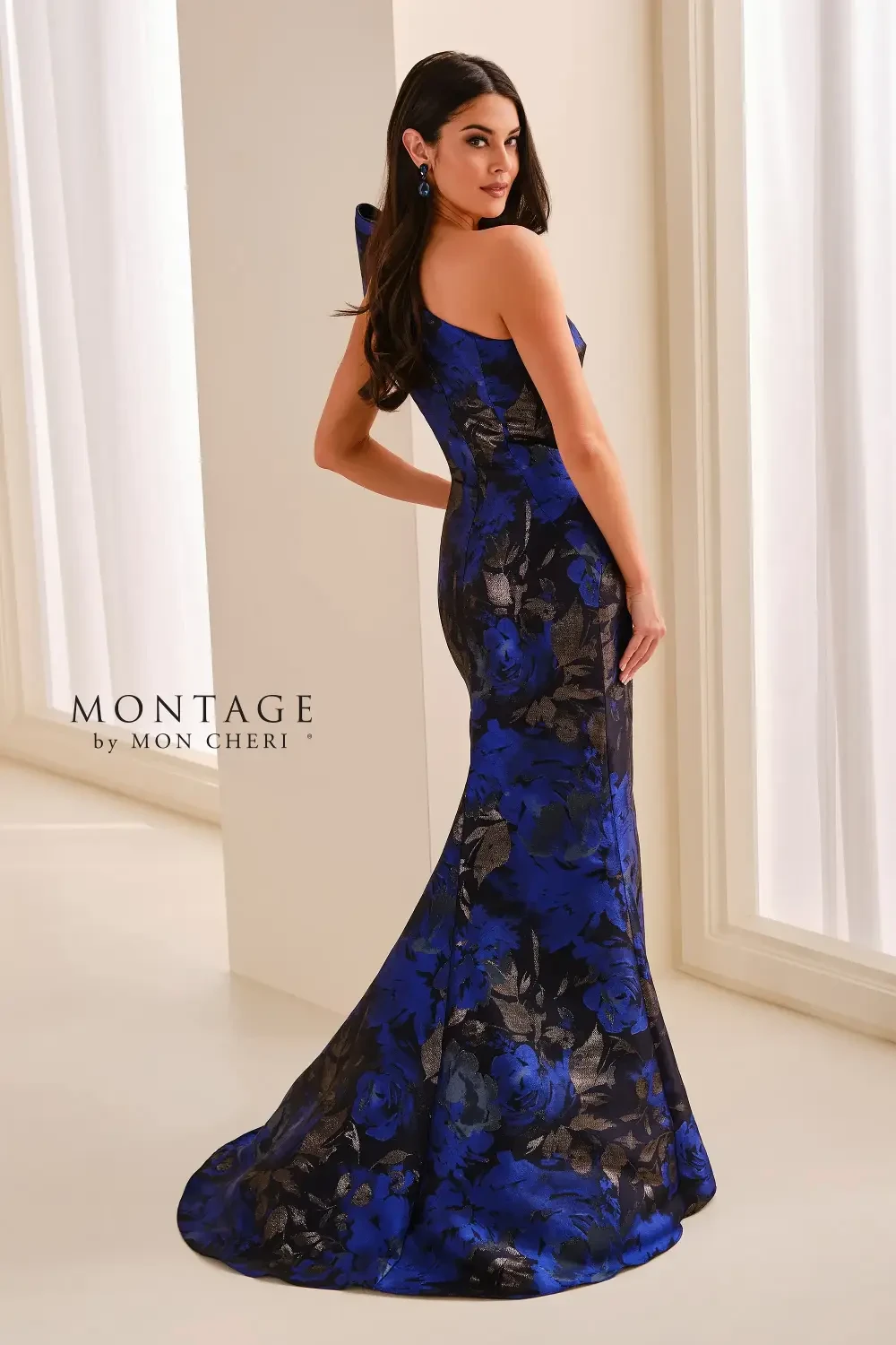 A woman wearing a blue and black floral evening gown with one shoulder and a large bow detail, posed elegantly in a light-colored room.