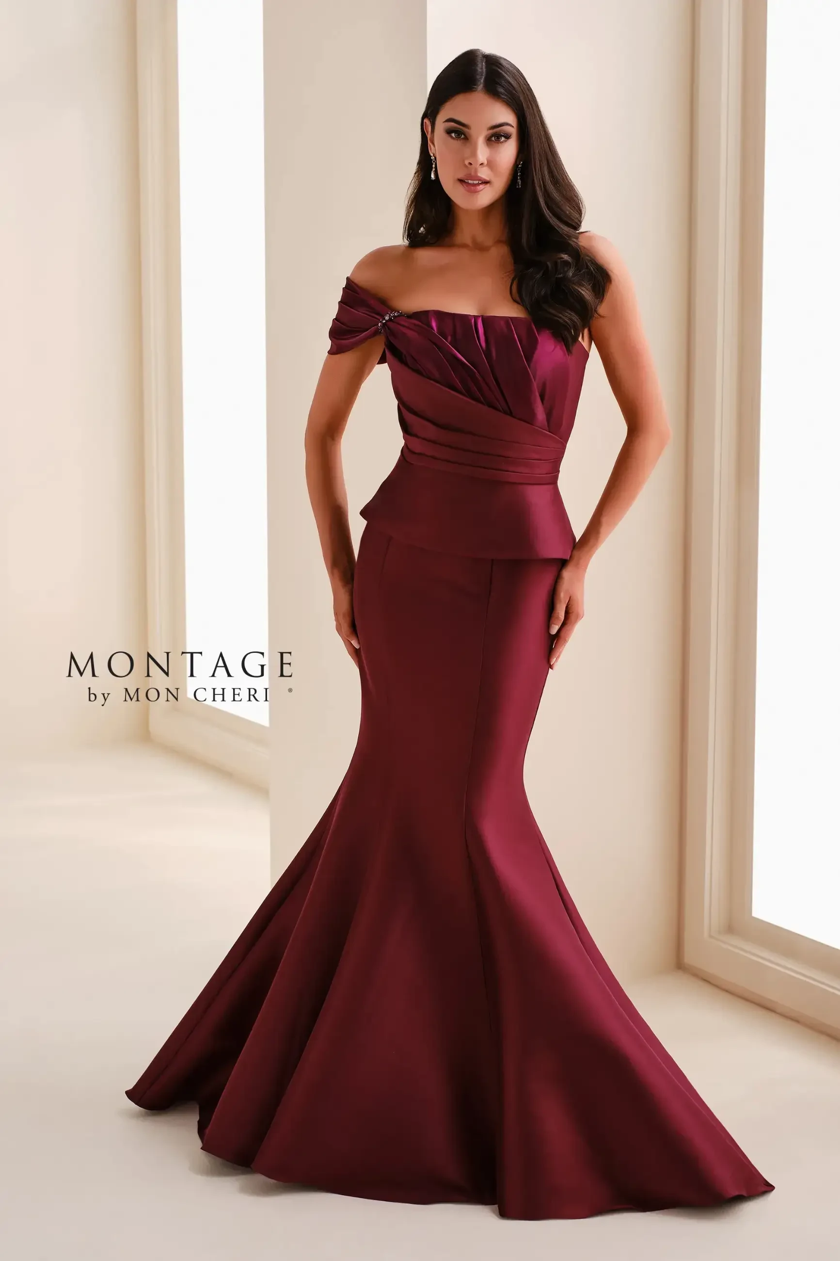 Woman in a strapless, mermaid-style burgundy gown with a fitted bodice and elegant pleats, posing confidently in a bright, minimalist room.