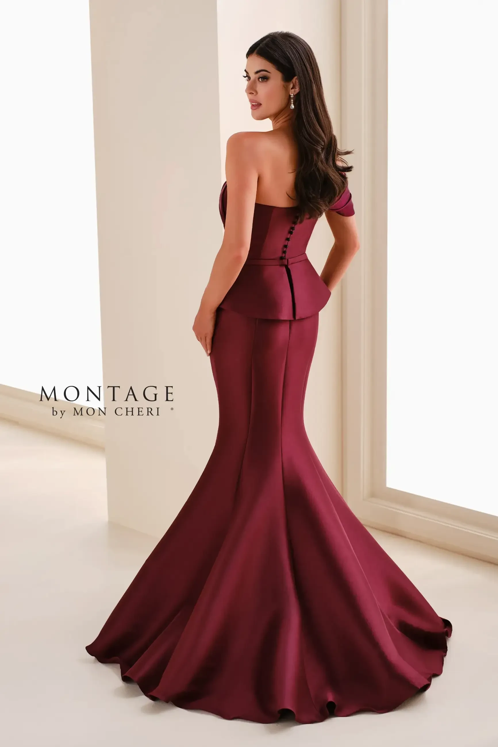 A woman in a burgundy, off-the-shoulder gown stands gracefully, facing a window. The dress features a fitted bodice and mermaid-style skirt. Elegant and sophisticated.