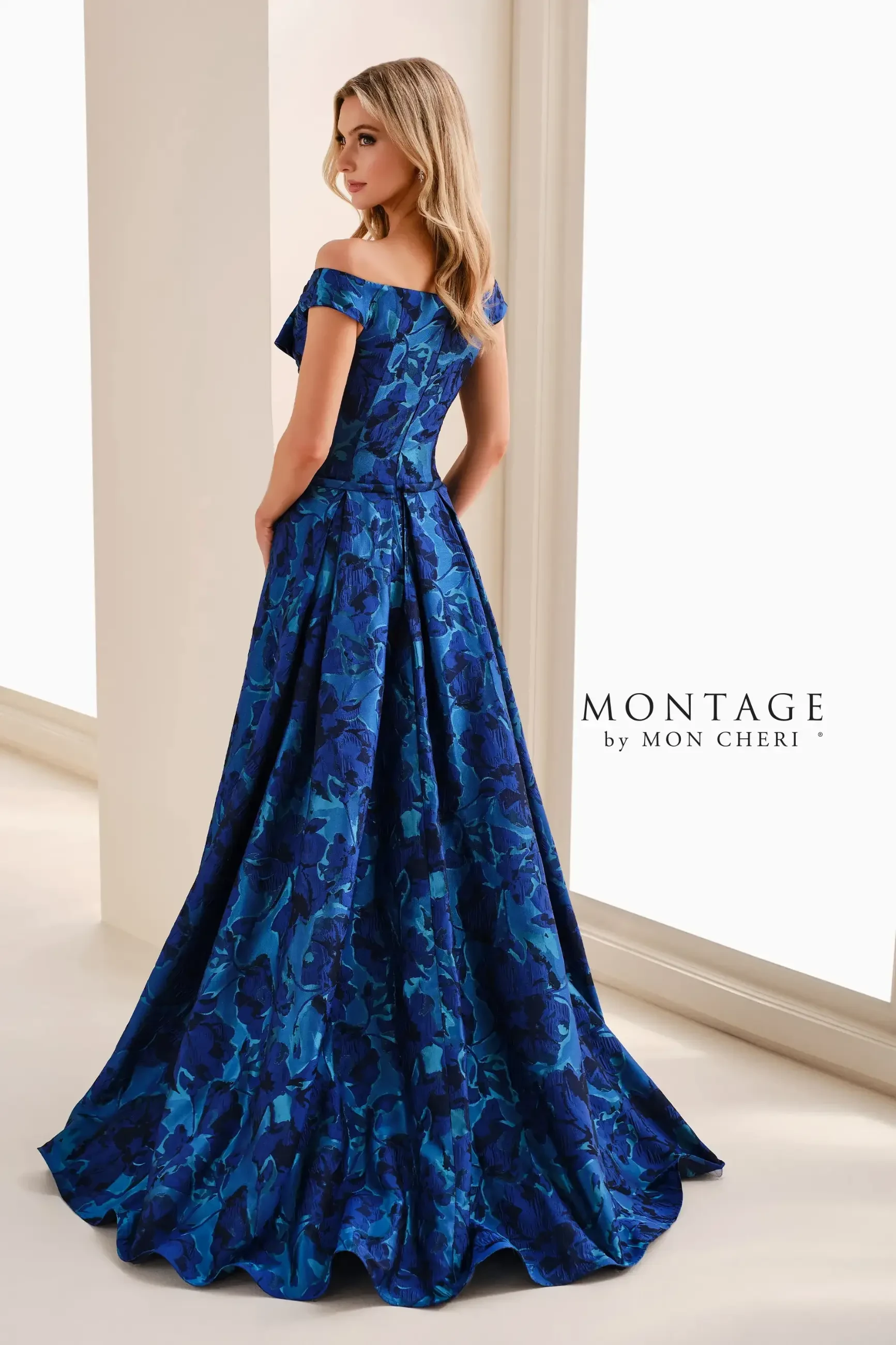 A woman in an elegant blue off-the-shoulder gown with floral patterns stands near a bright window. The dress flares out gracefully, exuding sophistication.