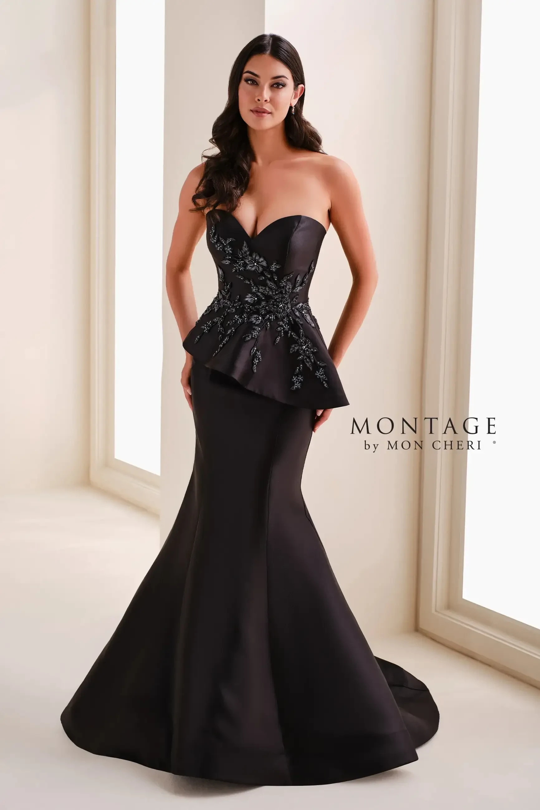 A woman wears an elegant, strapless black gown with floral embroidery and a flared skirt. She stands confidently in a softly lit room.